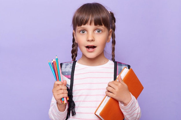 Foto interior de estudante surpresa carregando livros e lápis posando isolado sobre fundo roxo de volta à escola olhando para a câmera com olhos grandes