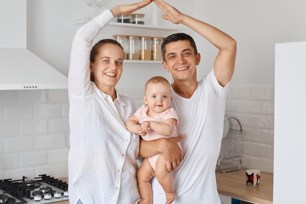 Foto interior de feliz casal sorridente em pé com a filha bebê e fazendo telhado com as mãos acima de suas cabeças se sente em segurança juntos em casa expressando emoções positivas