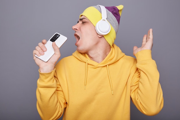 Foto interior de homem alegre e satisfeito com capuz amarelo cantando música e usando telefone inteligente como microfone se divertindo usando fone de ouvido sem fio dançando isolado sobre fundo cinza