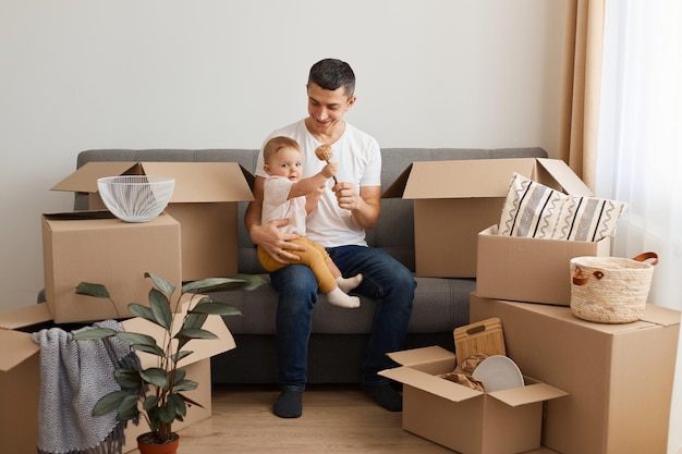 Foto interior de homem feliz sorridente, vestindo camiseta branca e jeans, sentado no sofá com a filha bebê cercada de caixas de papelão, brincando com o bebê enquanto desembala os pertences após a realocação.