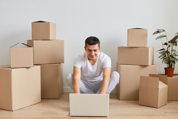 Foto interior de homem moreno bonito vestindo roupas casuais, sentado no chão perto de pacotes de papelão e trabalhando no laptop, terminando o projeto on-line enquanto se muda para um novo apartamento