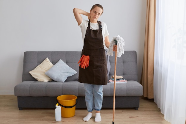 Foto interior de jovem cansada de limpar a casa descansando perto do sofá depois de lavar o chão com esfregão sentindo dor no pescoço tendo expressão exausta