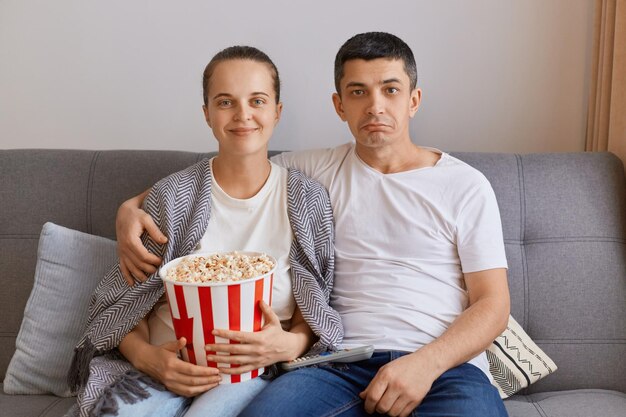 Foto interior de jovem casal assistindo tv em casa sentado na tosse abraçando a esposa embrulhada em manta e segurando balde com família de pipoca passando tempo juntos