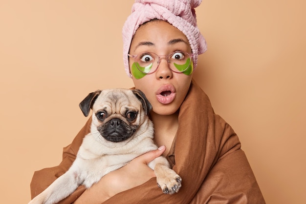 Foto interior de jovem chocada encara olhos esbugalhados mantém a boca aberta poses com cachorro pug favorito usa toalha de banho na cabeça e cobertor isolado sobre fundo marrom reage a notícias chocantes