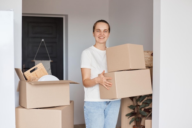 Foto interior de jovem se mudando para novo apartamento segurando caixas de papelão com pertences, olhando para a câmera com um sorriso feliz, desempacotando a pilha pessoal.