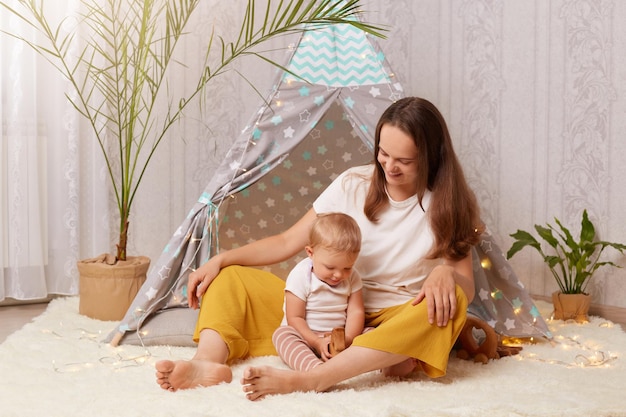 Foto interior de mãe alegre sorridente, vestindo camiseta branca sentada na tenda e olhando para seu filho infantil brincando com brinquedo de madeira