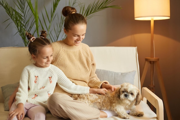 Foto interior de mãe e filha brincando com cachorro no sofá em casa menina feliz com cachorro pequinês e mãe relaxando em casa família com animal de estimação se divertindo juntos