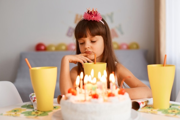 Foto interior de menina triste entediada com flor no cabelo sentada à mesa com bolo de aniversário linda garota com expressão facial chateada expressando tristeza para comemorar a festa sozinha