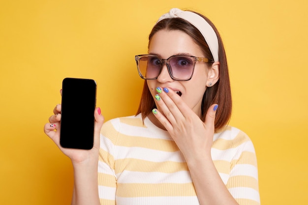 Foto interior de mulher caucasiana vestindo camisa listrada posando isolada sobre fundo amarelo mostrando celular com tela vazia com espaço para promoção