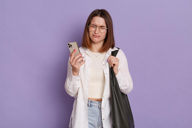 Foto interior de mulher deprimida triste com bolsa preta vestindo camisa branca e jeans posando isolado sobre fundo roxo em pé com o celular nas mãos e chorando com más notícias