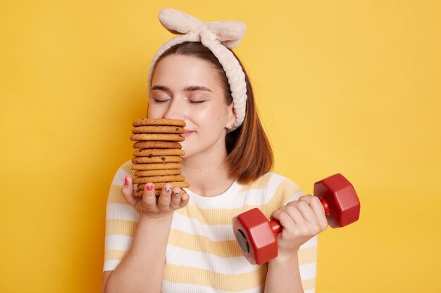 Foto interior de mulher encantada satisfeita vestindo camisa listrada e faixa de cabelo segurando haltere vermelho e cheirando biscoitos posando isolado sobre fundo amarelo como comer doces e malhar