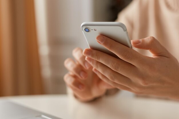 Foto interior de mulher sem rosto desconhecida vestindo suéter bege estilo casual usando telefone celular para verificar redes sociais ou digitar mensagens, furando o telefone inteligente nas mãos.