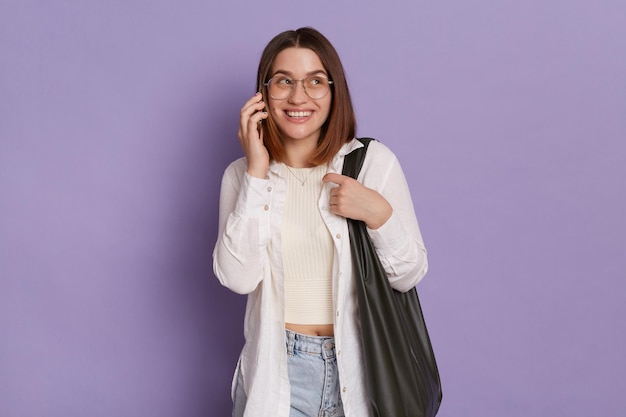 Foto interior de mulher sonhadora sorridente vestindo roupas elegantes segurando bolsa posando isolada sobre fundo roxo falando telefone olhando para longe com sorriso