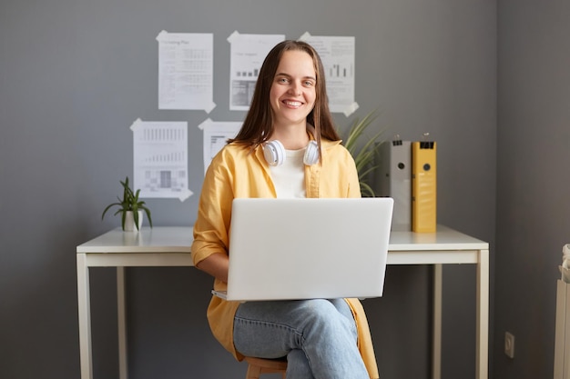 Foto interior de mulher sorridente freelancer ou estudante vestindo jaqueta amarela sentada no local de trabalho e trabalhando no laptop olhando para a câmera com sorriso expressando emoções positivas