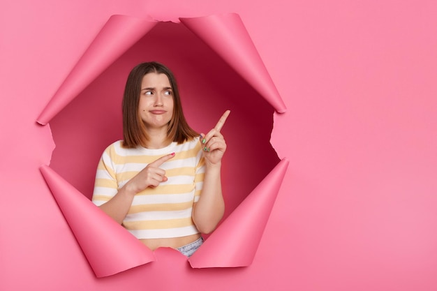 Foto interior de mulher triste chateada com lábios de beicinho vestindo camisa listrada, olhando através do avanço do fundo rosa apresentando área de publicidade apontando os dedos para longe