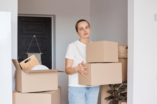 Foto interior de mulher triste vestindo camiseta branca estilo casual e jeans em pé com caixas de papelão com coisas, olhando para a câmera, expressando tristeza, ficando chateada por sair de casa.