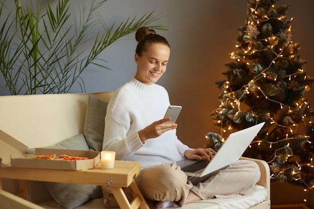 Foto interior de mulher usando smartphone enquanto trabalhava laptop em casa no Natal sentado no sofá perto da árvore de Natal com luzes lendo mensagem sms no celular durante as férias de inverno