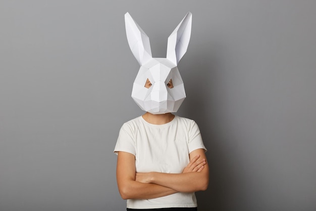 Foto interior de mulher vestindo camiseta branca e máscara de coelho de papel posando isolada sobre fundo cinza em pé calmo com os braços cruzados olhando para a câmera