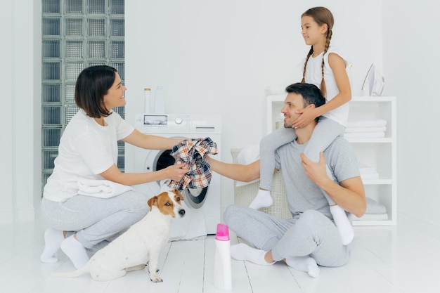 Foto interior de pai afetuoso brinca com filha na sala de lavagem dá camisa suja quadriculada para esposa posar juntos perto de máquina de lavar cercada com móveis brancos de garrafa de sabão em pó