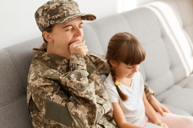 Foto interior de soldado jovem adulto sonolento sentado no sofá com sua filha e bocejando cansado depois de um longo caminho mãe voltando para casa da reunião da família do exército após longa despedida
