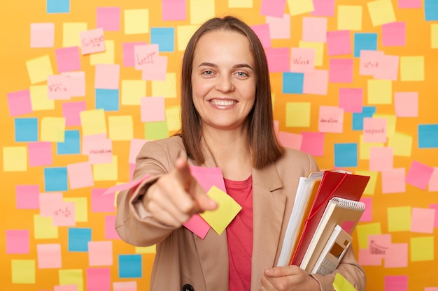 Foto interior de sorridente feliz alegre mulher caucasiana vestindo jaqueta bege empresária com documentos apontando para você escolhendo você para o trabalho posando contra a parede amarela com cartões de memorando coloridos