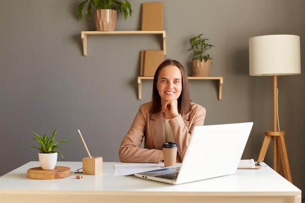 Foto interior de sorridente freelancer ou gerente de mulher caucasiana satisfeita vestindo jaqueta bege sentada à mesa perto do laptop olhando para a câmera segurando o queixo