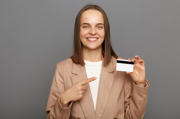 Foto foto interior de sorridente mulher de cabelos castanhos satisfeita vestindo jaqueta apontando para cartão de crédito recomendar banco ou descontos de compras isolados sobre fundo cinza