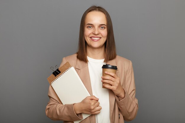 Foto interior de sorridente mulher de cabelos castanhos satisfeita vestindo jaqueta segurando prancheta com documentos e copo descartável com café isolado sobre fundo cinza