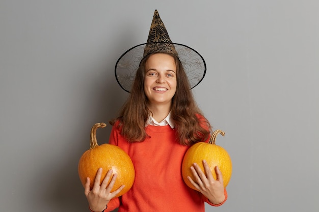 Foto interior de sorridente mulher encantada com chapéu de bruxa em pé com duas abóboras laranja isoladas sobre fundo cinza celebrando o Halloween olhando para a câmera com um sorriso