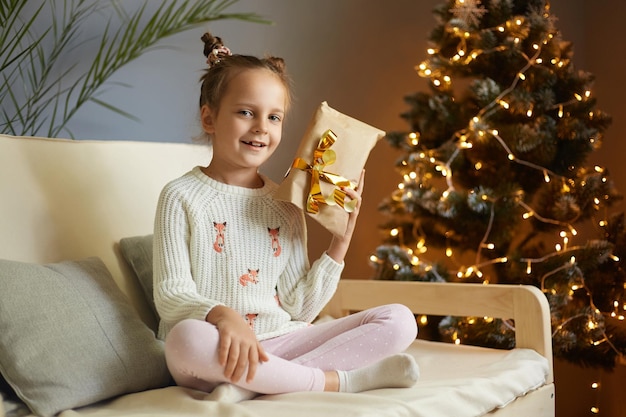 Foto interior de uma adorável garotinha bonita com dois pães de cabelo vestindo roupas casuais sentadas no sofá e mostrando seu presente de Natal para a câmera