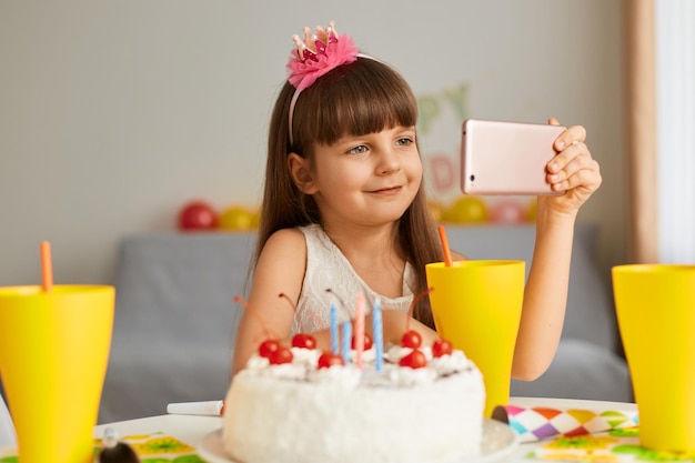 Foto interior de uma garotinha encantadora comemorando seu aniversário, segurando o telefone inteligente nas mãos e fazendo videochamada com fridens, mostrando seu bolo, sorrindo.