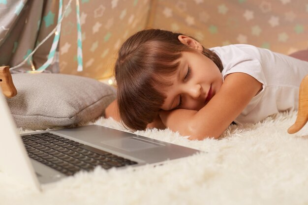 Foto interior de uma garotinha sonolenta de cabelos escuros vestindo camiseta branca ou pijama deitada no chão em wigwam com os olhos fechados adormecendo enquanto assiste a desenhos na tenda peetee