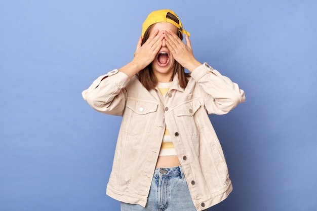 Foto foto interior de uma jovem adolescente animada usando boné de beisebol e jaqueta cobrindo os olhos com as palmas das mãos gritando mantém a boca aberta posando isolada sobre fundo azul