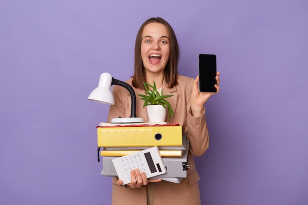 Foto interior de uma jovem adulta alegre espantada vestindo jaqueta bege segurando pastas de papel isoladas sobre fundo roxo mostrando telefone celular com espaço de cópia de exibição vazio