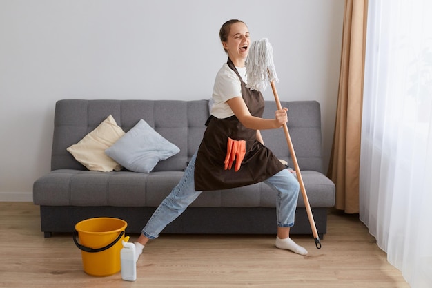 Foto interior de uma jovem excitada vestindo jeans e avental marrom limpando sua casa cantando no esfregão usando a ferramenta como microfone e se divertindo na sala de estar
