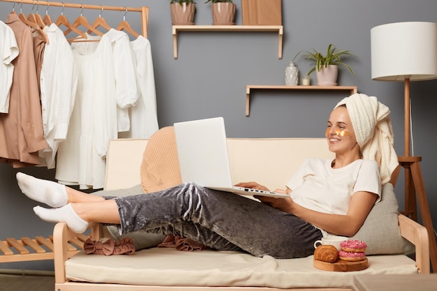 Foto interior de uma linda mulher sorridente vestindo calças brancas de camiseta cinza e com toalha na cabeça deitada na tosse com roupas penduradas na prateleira no fundo navegando na internet assistindo filmes