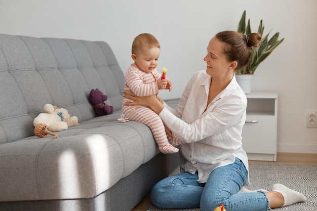 Foto interior de uma linda mulher vestindo camisa branca e jeans sentado no chão e brincando com o bebê sentado na tosse mãe passando tempo com sua filha expressando felicidade