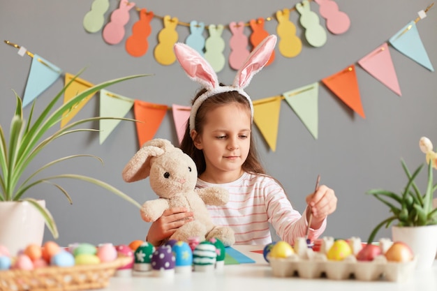 Foto interior de uma menina bonitinha e concentrada, vestida com orelhas de coelho, sentada à mesa e pintando ovos de Páscoa contra a parede cinza decorada, preparando-se para o feriado