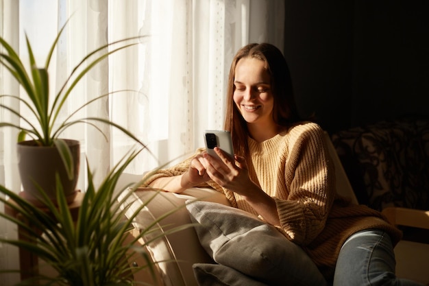 Foto interior de uma mulher alegre e relaxada sorridente sentada no sofá usando celular perto de uma grande janela aproveitando seu tempo de lazer conversando com amigos verificando redes sociais