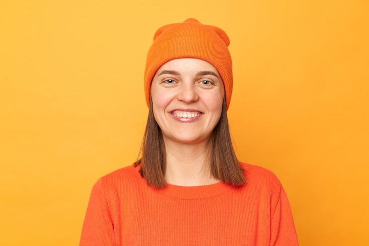 Foto foto interior de uma mulher bonita e atraente hipster vestindo suéter laranja e posando de chapéu isolado sobre fundo amarelo olhando para a câmera com sorriso de dentes expressando felicidade