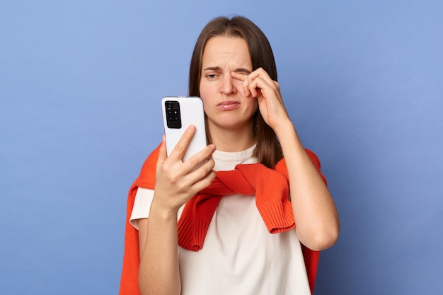 Foto interior de uma mulher cansada e triste vestindo camiseta branca e suéter laranja amarrado sobre os ombros em pé isolado no fundo azul usando telefone celular por longas horas sente dor nos olhos esfregando os olhos