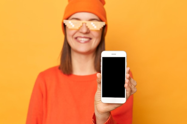 Foto foto interior de uma mulher caucasiana sorridente satisfeita usando gorro laranja e óculos no estilo minecraft em pé isolado sobre fundo amarelo mostrando telefone inteligente com tela vazia