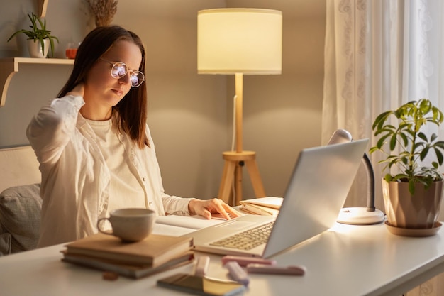 Foto interior de uma mulher exausta e cansada com cabelos castanhos sentada à mesa trabalhando no laptop à noite em casa sente fadiga e dor no pescoço massageando suas costas
