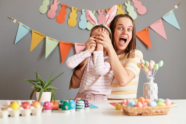 Foto interior de uma mulher maravilhada com o rosto surpreso cobrindo os olhos da filha com as mãos comemorando a Páscoa se divertindo em casa garotinha usando orelhas de coelho