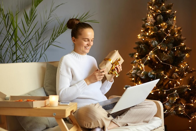 Foto interior de uma mulher satisfeita e encantada com penteado de coque usando um jumper branco sentado no sofá perto da árvore de Natal com videochamada no laptop mostrando a caixa de presente para o ano novo