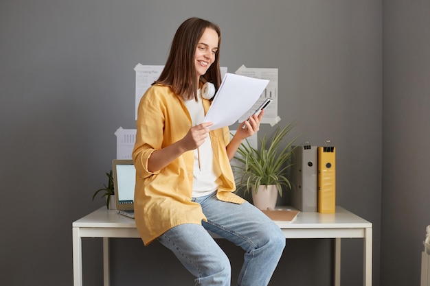 Foto interior de uma mulher sorridente atraente com cabelo castanho vestindo camisa amarela posando no escritório sentado na mesa e lendo informações em documentos sorrindo