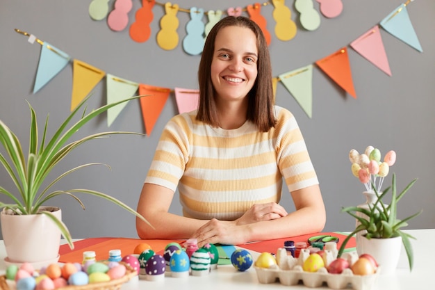 Foto interior de uma mulher sorridente atraente com cabelo castanho vestindo camisa listrada sentada à mesa pintando ovos de Páscoa em casa se preparando para as férias em casa no quarto decorado
