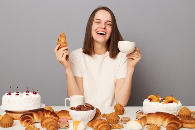 Foto interior de uma mulher sorridente e encantada com cabelos castanhos, sentada à mesa isolada sobre fundo cinza, bebendo chá com croissant, desfrutando de seu jantar rindo alegremente