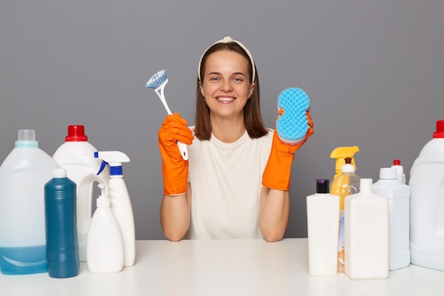 Foto interior de uma mulher sorridente feliz em camiseta branca e luvas laranja segurando escova e esponja para limpar a casa sendo fotografada entre diferentes detergentes posando isolado sobre fundo cinza