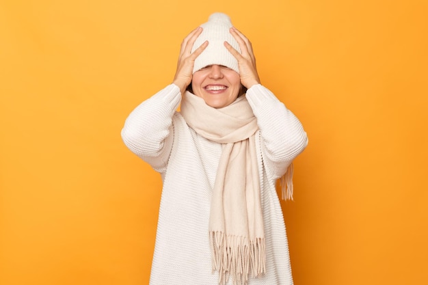 Foto interior de uma mulher sorridente satisfeita usando cachecol de suéter e chapéu escondendo os olhos com boné sorrindo alegremente se divertindo parece tímido em pé isolado sobre fundo amarelo